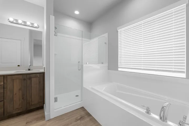 Soaking tub, walk-in shower with glass enclosure, wood-finish flooring, vanity with stone countertop, and recessed lighting