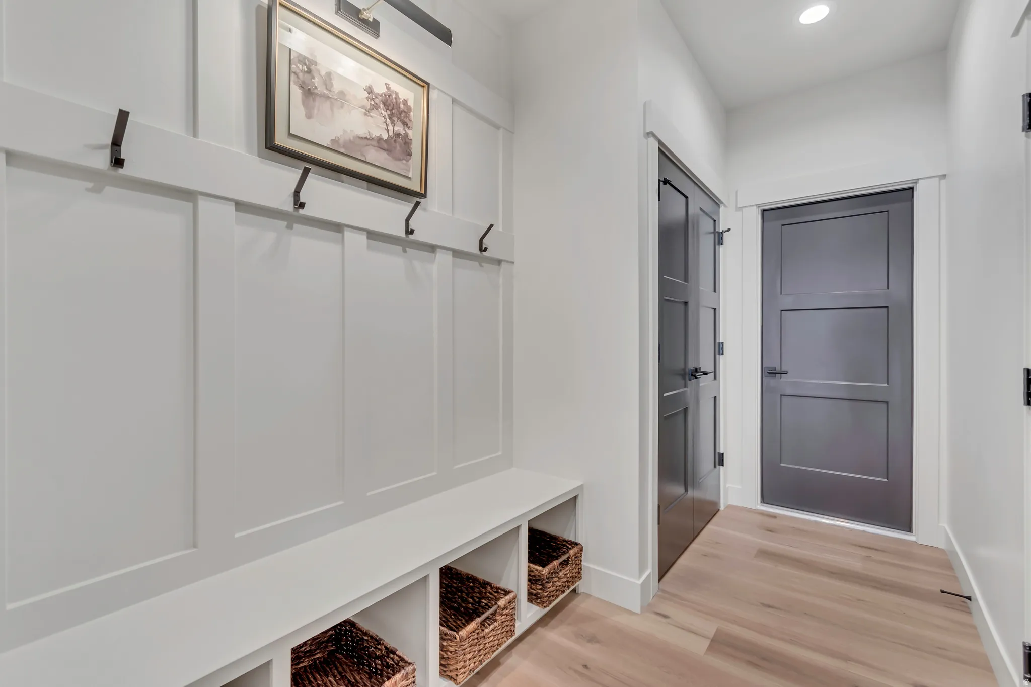 Built-in entryway bench with integrated storage cubbies, white board-and-batten wall treatment with dark metal hooks, light wood-finish flooring, and dark paneled interior doors