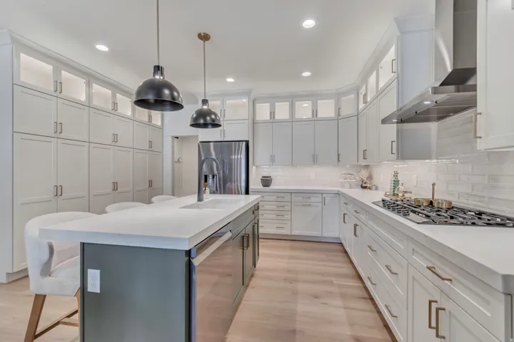 Expansive kitchen featuring abundant white cabinetry, light wood-finish flooring, a central island with integrated sink, a gas cooktop, and a stainless steel range hood