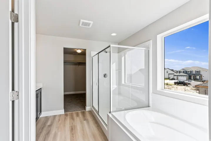 Soaking tub with a large window, glass-enclosed shower, wood-finish flooring, and a vanity with a light-colored countertop