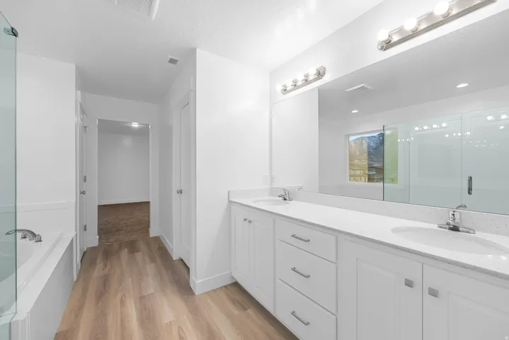 Double vanity with white cabinetry and integrated sinks, a large mirror, wood-finish flooring, and a glass-enclosed tub