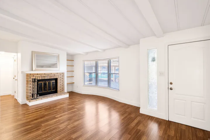 Wood-finish flooring, exposed beam ceiling, brick fireplace with metal insert, built-in shelving, and a bay window