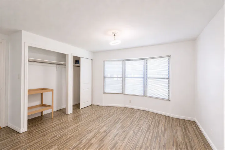 Spacious room featuring wood-finish flooring, white walls, and a bay window with blinds