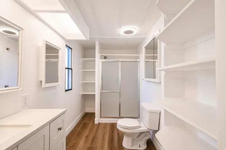Integrated vanity with a rectangular sink, wood-finish flooring, built-in shelving, glass-enclosed shower, and a large overhead light panel