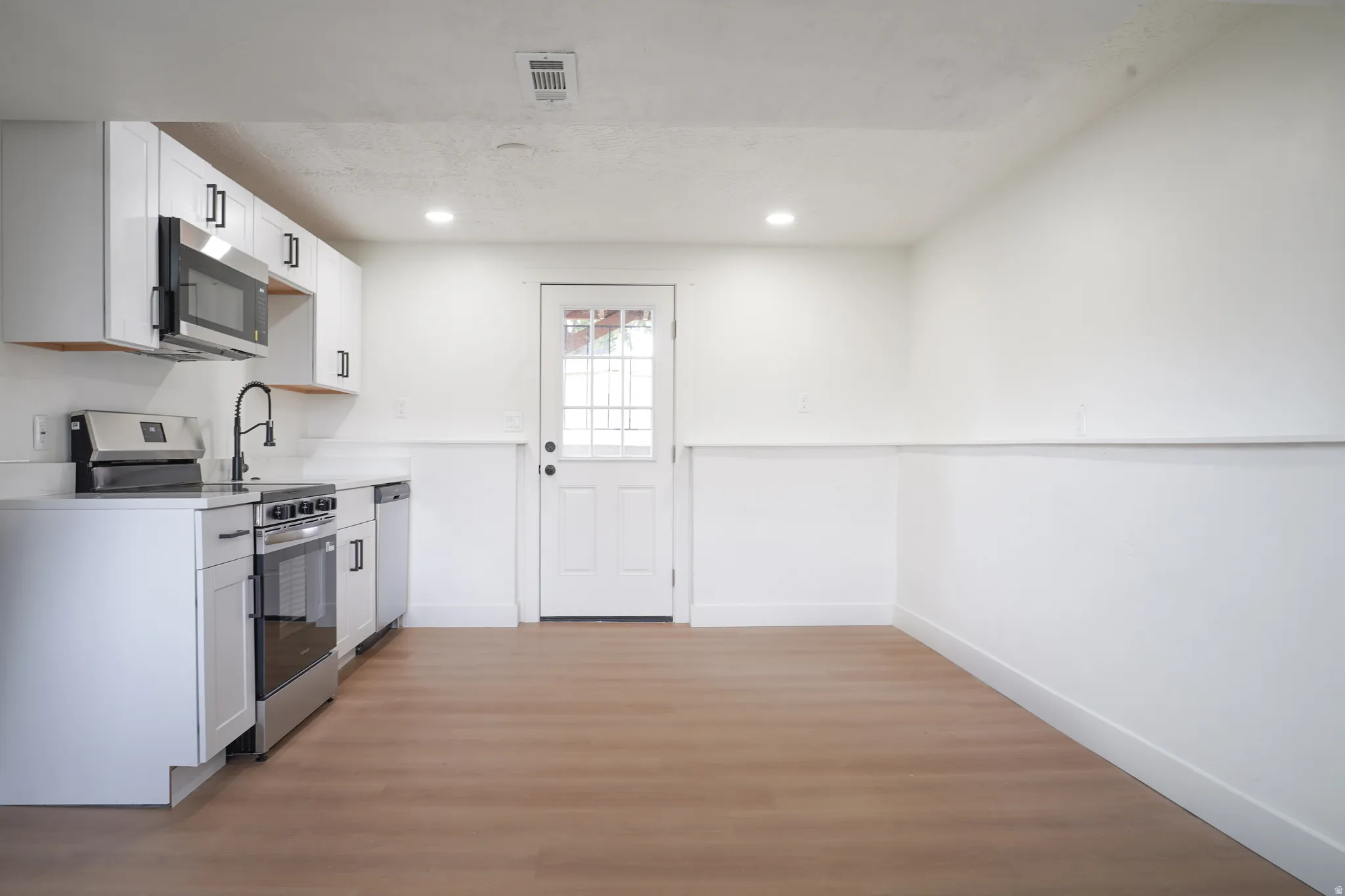White shaker cabinetry with matte black hardware, stainless steel appliances, wood-finish flooring, recessed lighting, and a paneled door with glass inserts