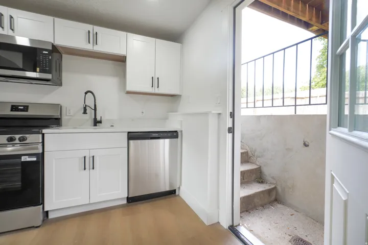 White shaker cabinetry with dark hardware, stainless steel appliances, light-toned countertops, wood-finish flooring, and a private exterior entrance