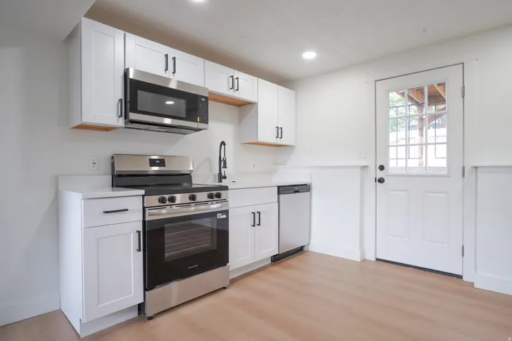 White shaker-style cabinetry with matte black hardware, stainless steel appliances, light wood-finish flooring, white countertops, and a white paneled door with window panes