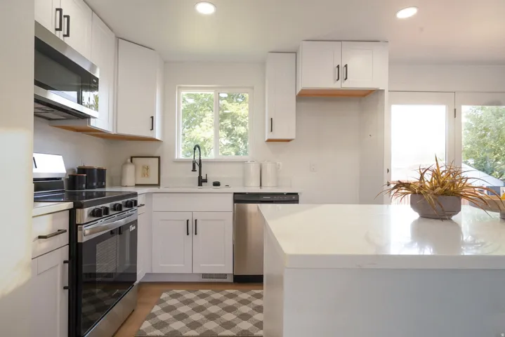 Modern kitchen featuring white shaker cabinetry, stainless steel appliances, a white solid surface countertop, wood-finish flooring, and a large window