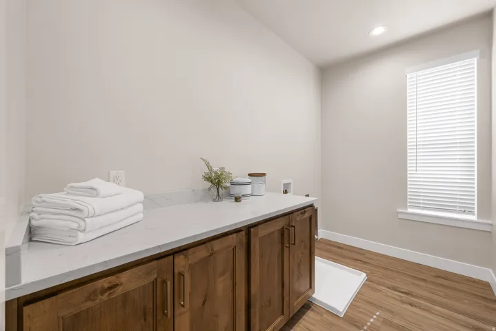 Wood-finish flooring, recessed ceiling light, wood cabinetry with bronze hardware, white stone countertop, and window with blinds