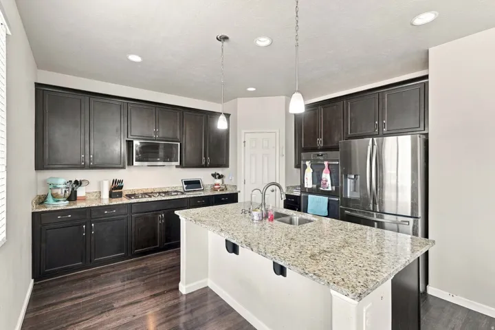 Kitchen featuring dark wood-finish cabinetry, light speckled countertops, stainless steel appliances, a center island with sink, and wood-finish flooring
