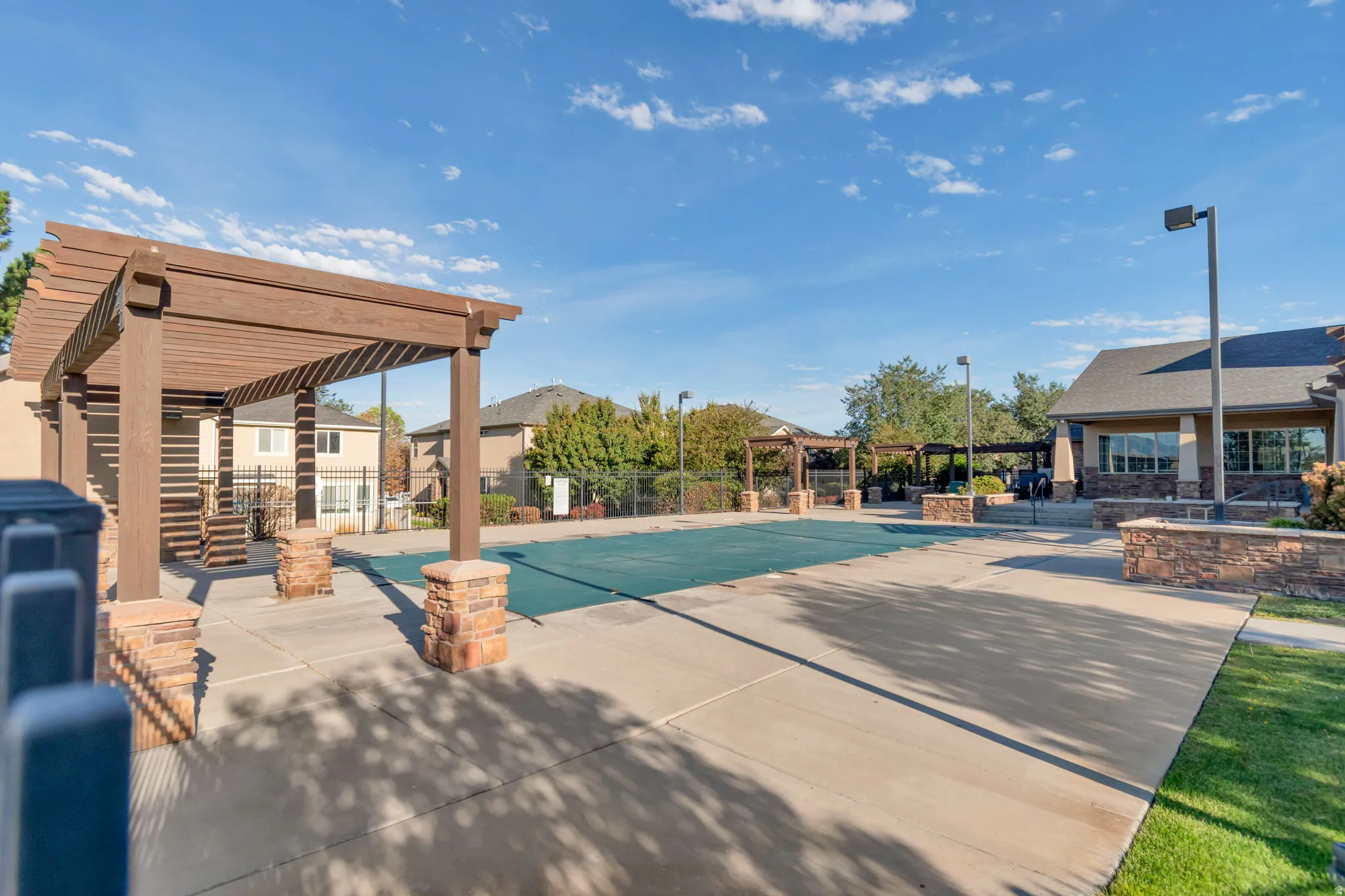 Community Pool.  Two wood pergolas with stone-clad bases, a pool with a cover, and a clubhouse with stone accents
