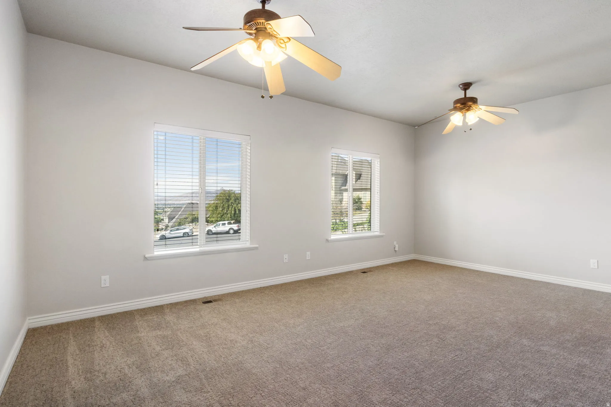 Spacious room with sloped ceilings, two ceiling fans with integrated lighting, two windows with blinds, and neutral carpeting