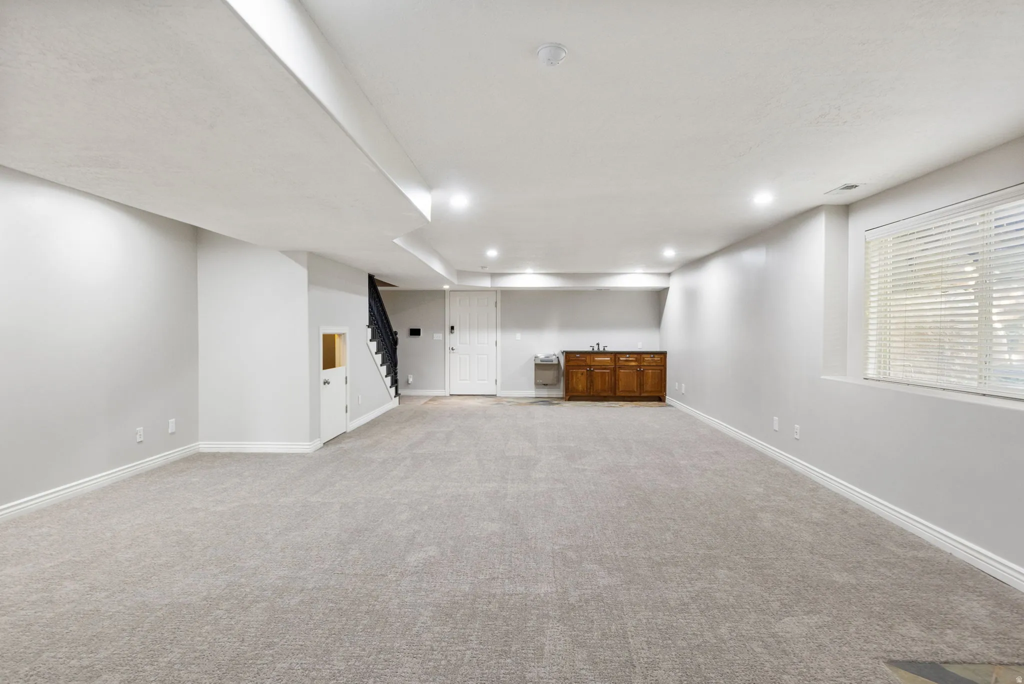 Expansive carpeted area with recessed lighting, built-in wooden cabinetry, sink, and a window with blinds