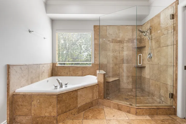 Tiled shower with a built-in bench, corner soaking tub with deck-mounted fixtures, and a window with blinds