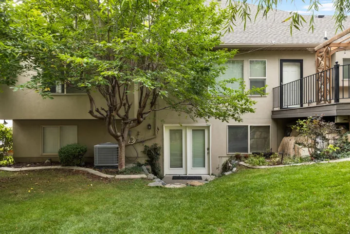 Manicured lawn with mature tree, lower-level double doors, upper-level balcony with black railing, and stucco exterior
