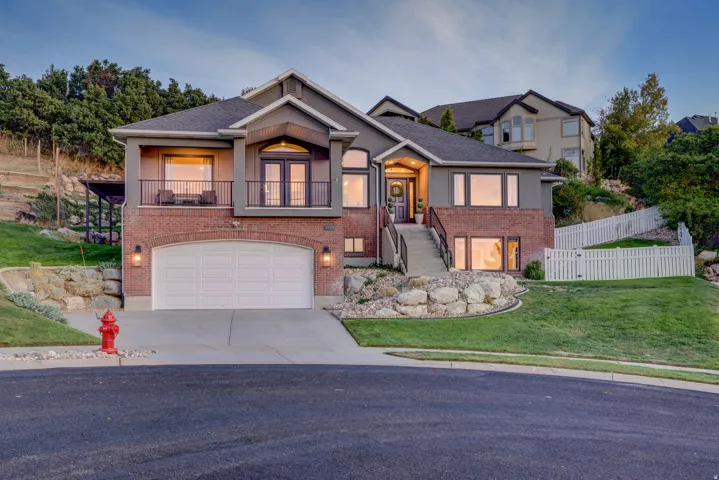 Two-story residence featuring a brick and stucco exterior, an arched garage entrance, a prominent front balcony with double doors, and a white picket fence enclosing a sloped lawn