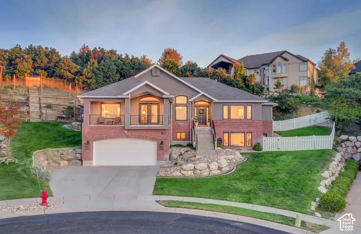 Multi-level exterior featuring a brick and siding facade, an attached garage, a covered balcony with metal railing, tiered rock retaining walls, and white vinyl privacy fencing
