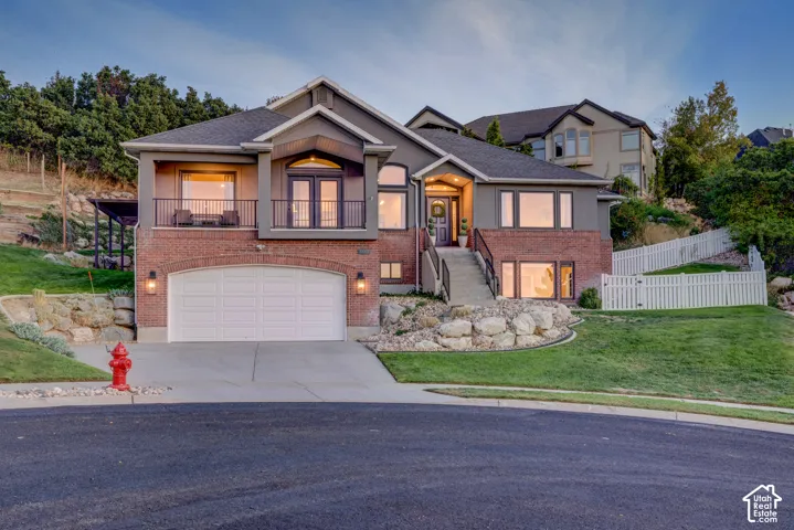 Two-story residence featuring a brick and stucco exterior, an arched garage entrance, a prominent front balcony with double doors, and a white picket fence enclosing a sloped lawn
