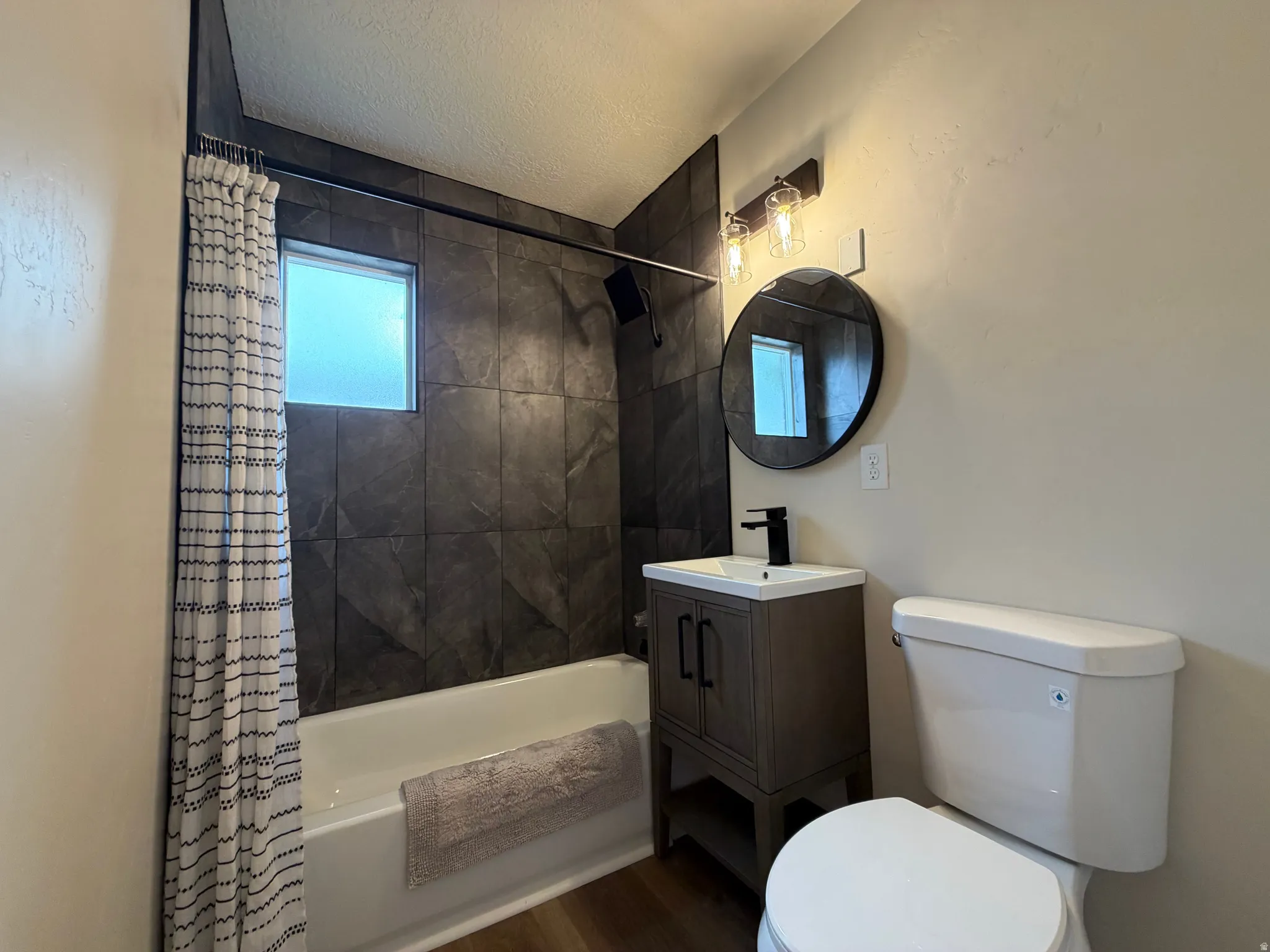 Bathtub with dark-toned wall tile, wood-finish flooring, single vanity with a black faucet, and a round mirror