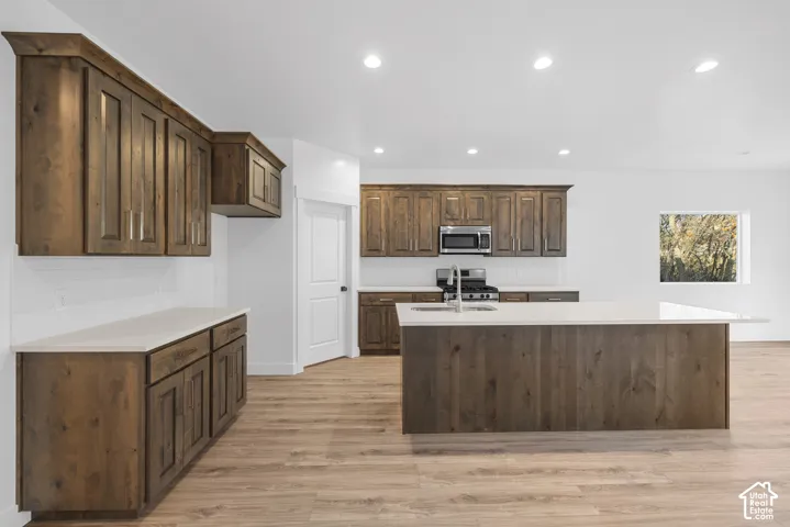 Kitchen featuring wood-finish cabinetry, light-toned countertops, a central island with a sink, stainless steel appliances, and recessed lighting