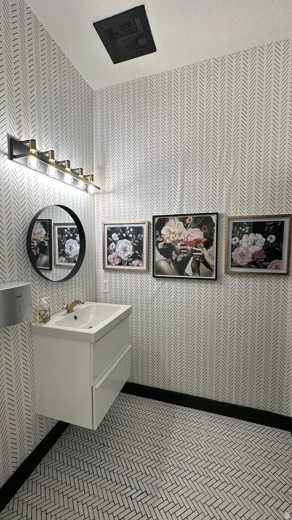 Floating vanity with integrated sink, patterned wallpaper, wall-mounted light fixture, circular mirror, and herringbone tile flooring