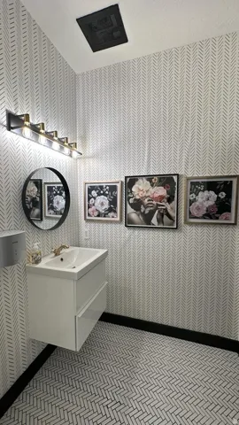 Floating vanity with integrated sink, patterned wallpaper, wall-mounted light fixture, circular mirror, and herringbone tile flooring