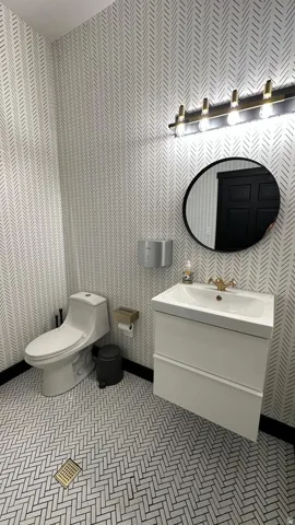 Herringbone pattern tile flooring, wall-mounted vanity with integrated sink, gold-finish faucet, contemporary vanity light fixture, and patterned wallpaper