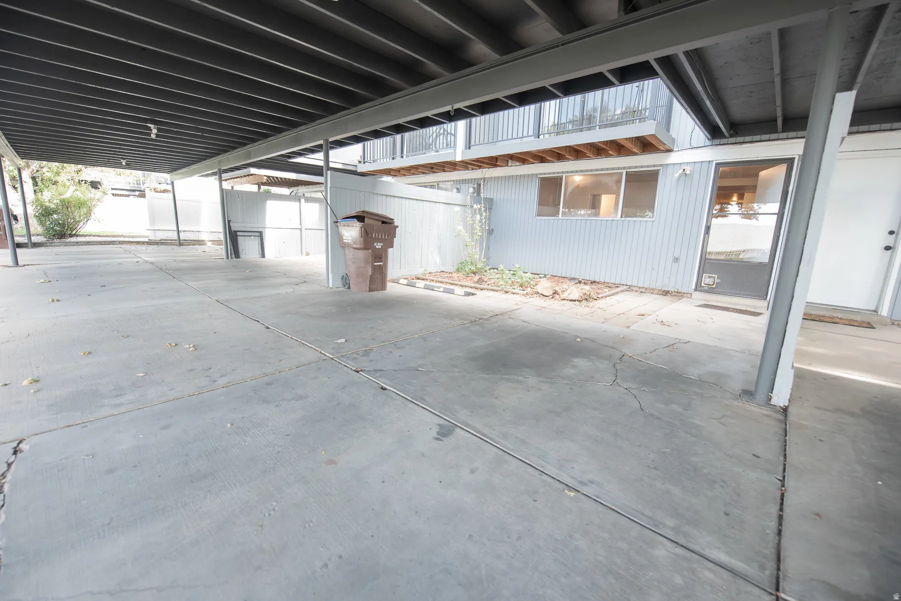 Covered parking area with concrete flooring, framed by structural supports, and a distinct exterior wall featuring windows and an entry door