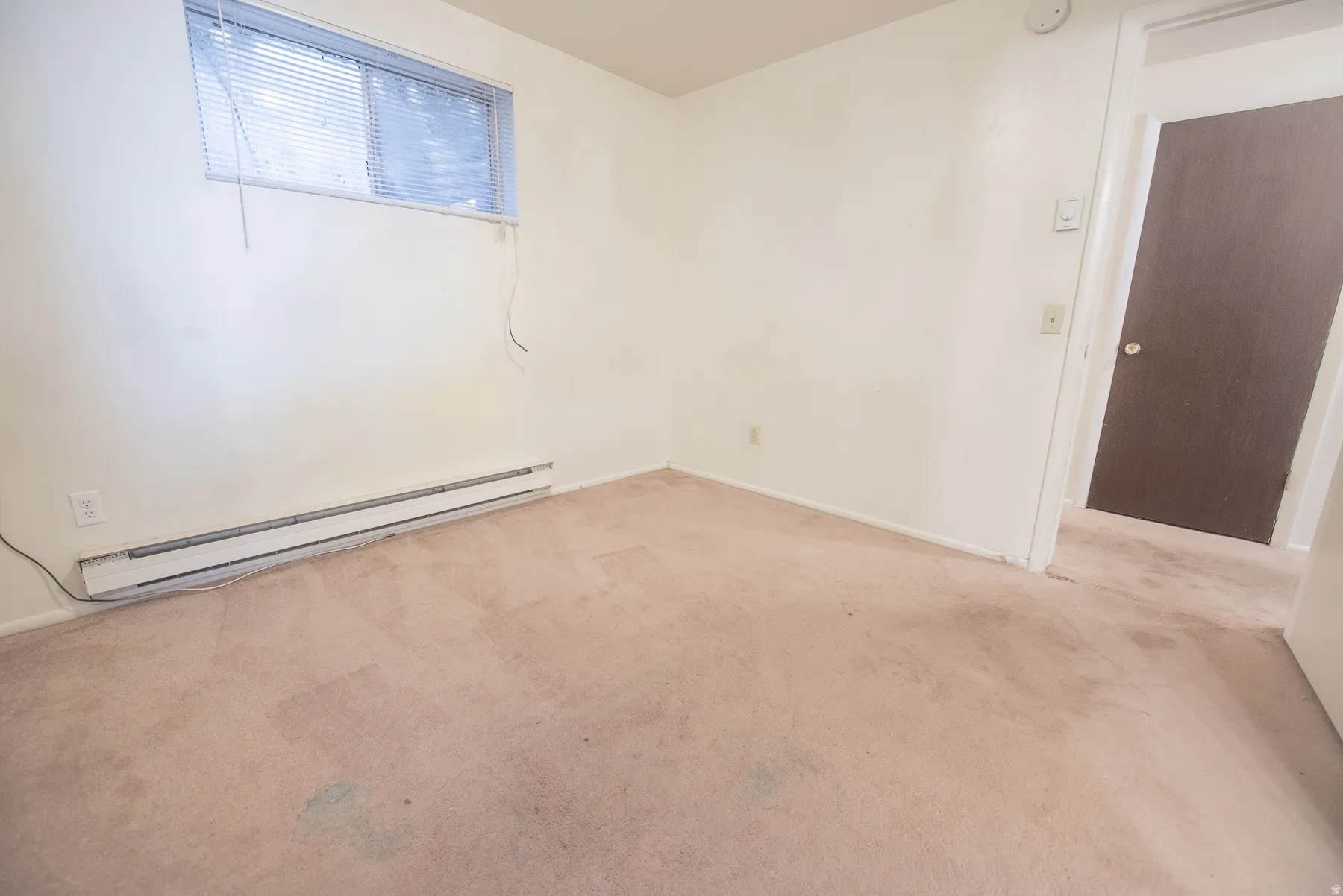 Neutral carpet flooring, white painted walls, horizontal blinds, baseboard heater, and a brown interior door