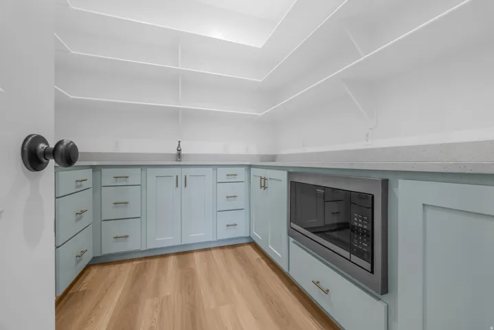 Built-in microwave, wood-finish flooring, extensive white shelving, light blue cabinetry with gold-tone hardware, and light-toned countertops