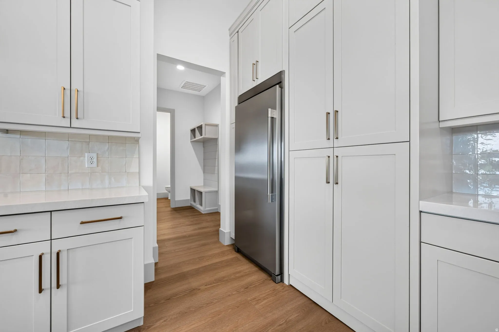 White shaker-style cabinetry, stainless steel refrigerator, light-toned countertop, square tile backsplash, and wood-finish flooring