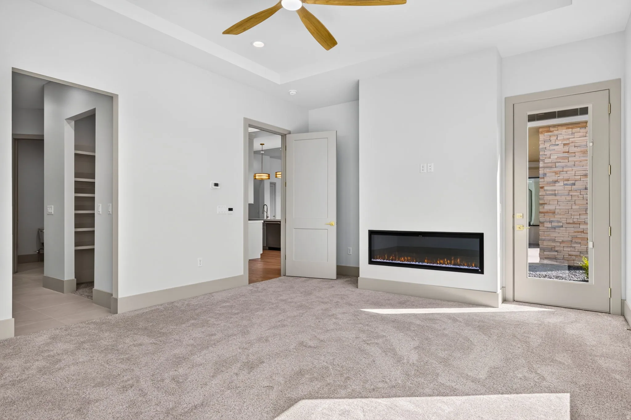 Recessed ceiling with an overhead fan, built-in electric fireplace, light-toned carpeting, built-in shelving, and a glass-paneled door