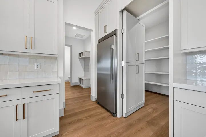 White shaker cabinetry with bronze hardware, white countertops, tile backsplash, wood-finish flooring, and a walk-in pantry with shelving