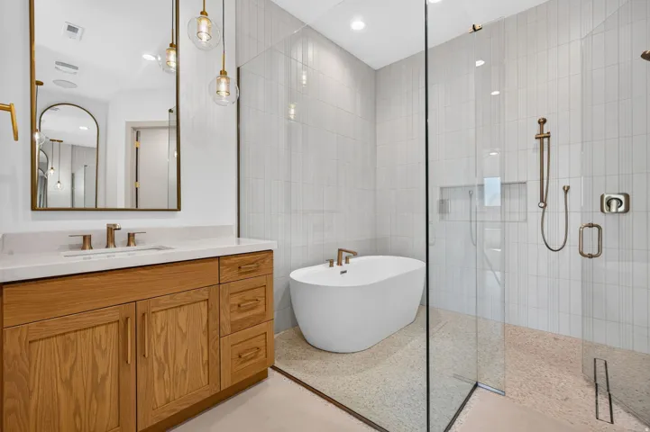 Freestanding soaking tub, glass-enclosed shower, wood-finish vanity, light-toned countertop, and vertical tile shower surround
