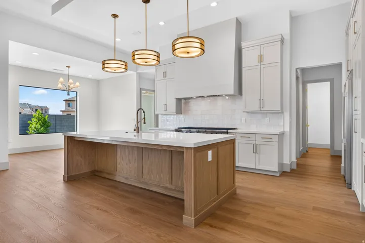 Spacious kitchen featuring a large island with a light-toned countertop, wood-finish flooring, white cabinetry with gold-toned hardware, and a tile backsplash