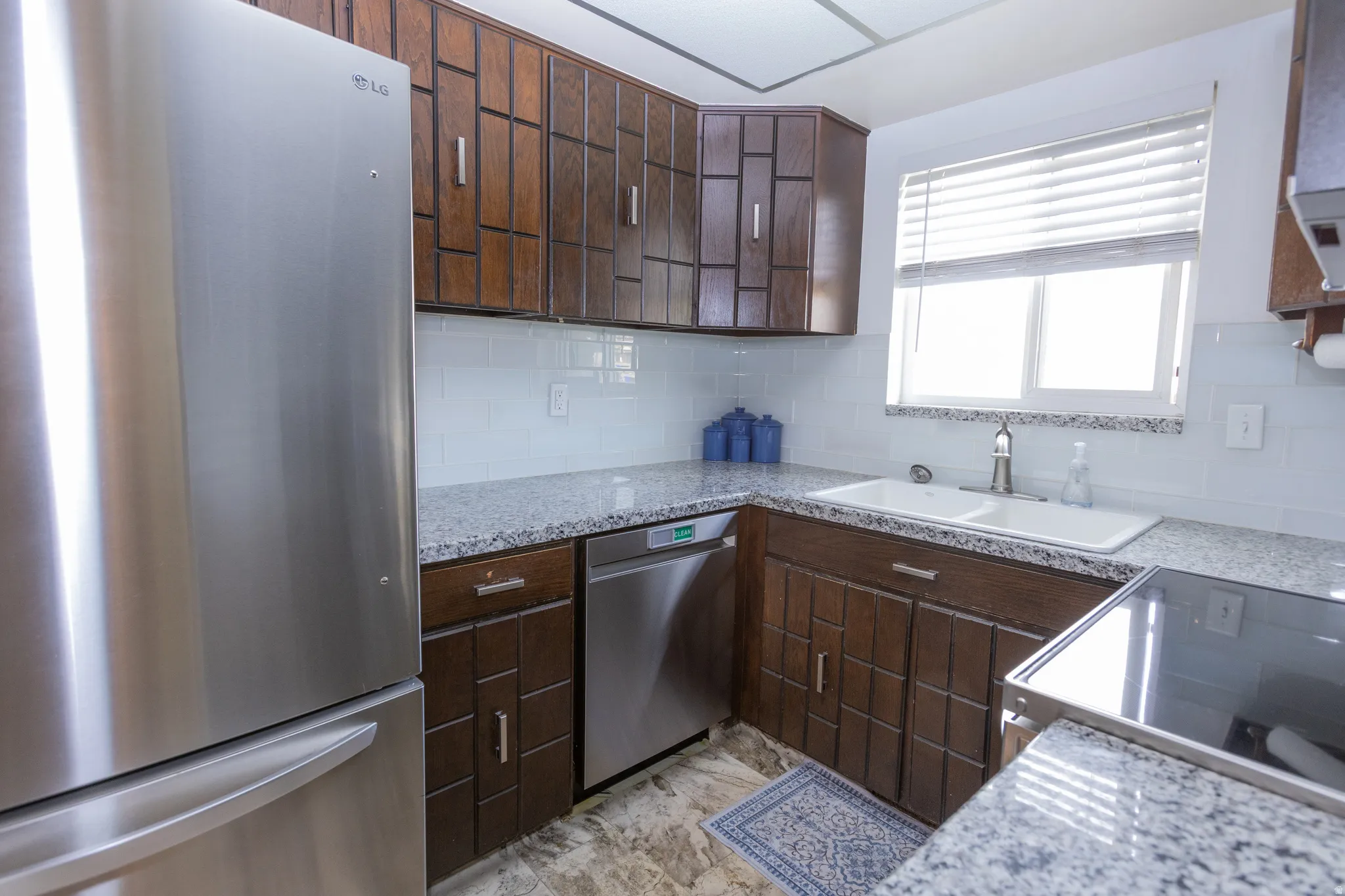 Stainless steel refrigerator, granite-style countertops, subway tile backsplash, dark wood-finish cabinetry, and a dual basin sink