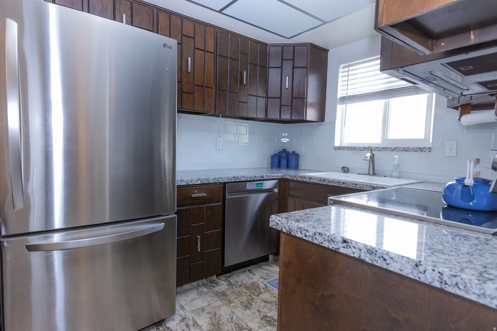Stainless steel refrigerator, granite countertops, white subway tile backsplash, dark wood cabinetry, and stainless steel dishwasher