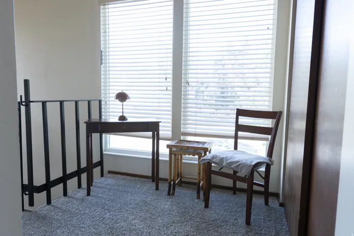 Carpeted floor with a large window, horizontal blinds, and a built-in wood panel