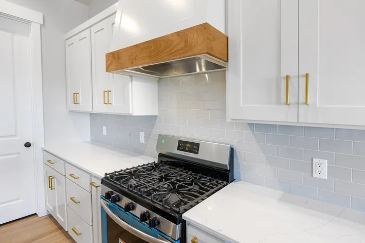 White shaker-style cabinetry with gold-tone hardware, a stainless steel gas range, light-toned countertops, a light subway tile backsplash, and a wood-finish range hood accent