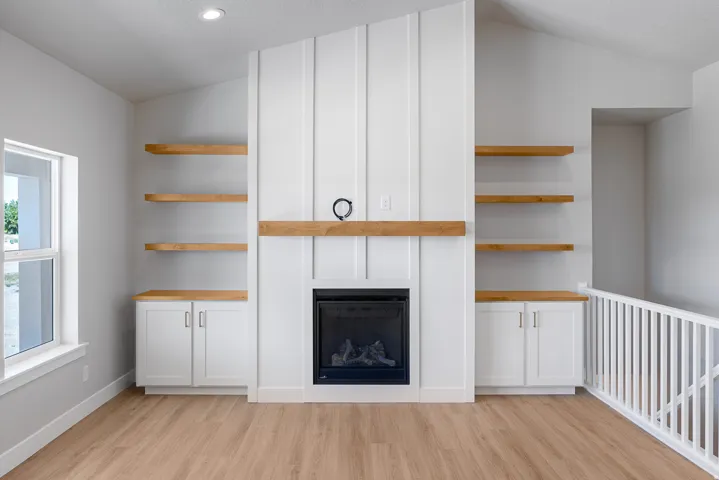 Vaulted ceiling, wood-finish flooring, built-in cabinetry with wood-finish tops, electric fireplace with wood mantel, and integrated shelving