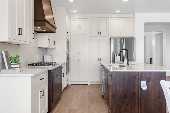 White cabinetry with dark contrasting island cabinetry, stainless steel appliances, solid surface countertops, wood-finish flooring, and a gas range with a decorative wood hood