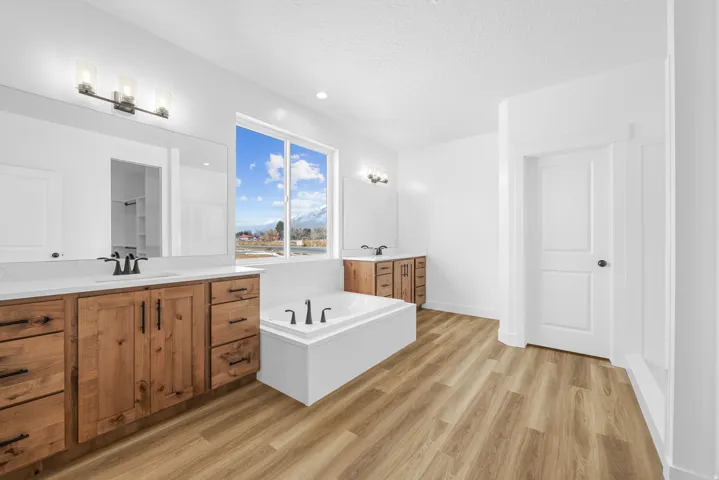 Dual wood-finish vanities with white countertops, a built-in soaking tub, wood-finish flooring, and a large window