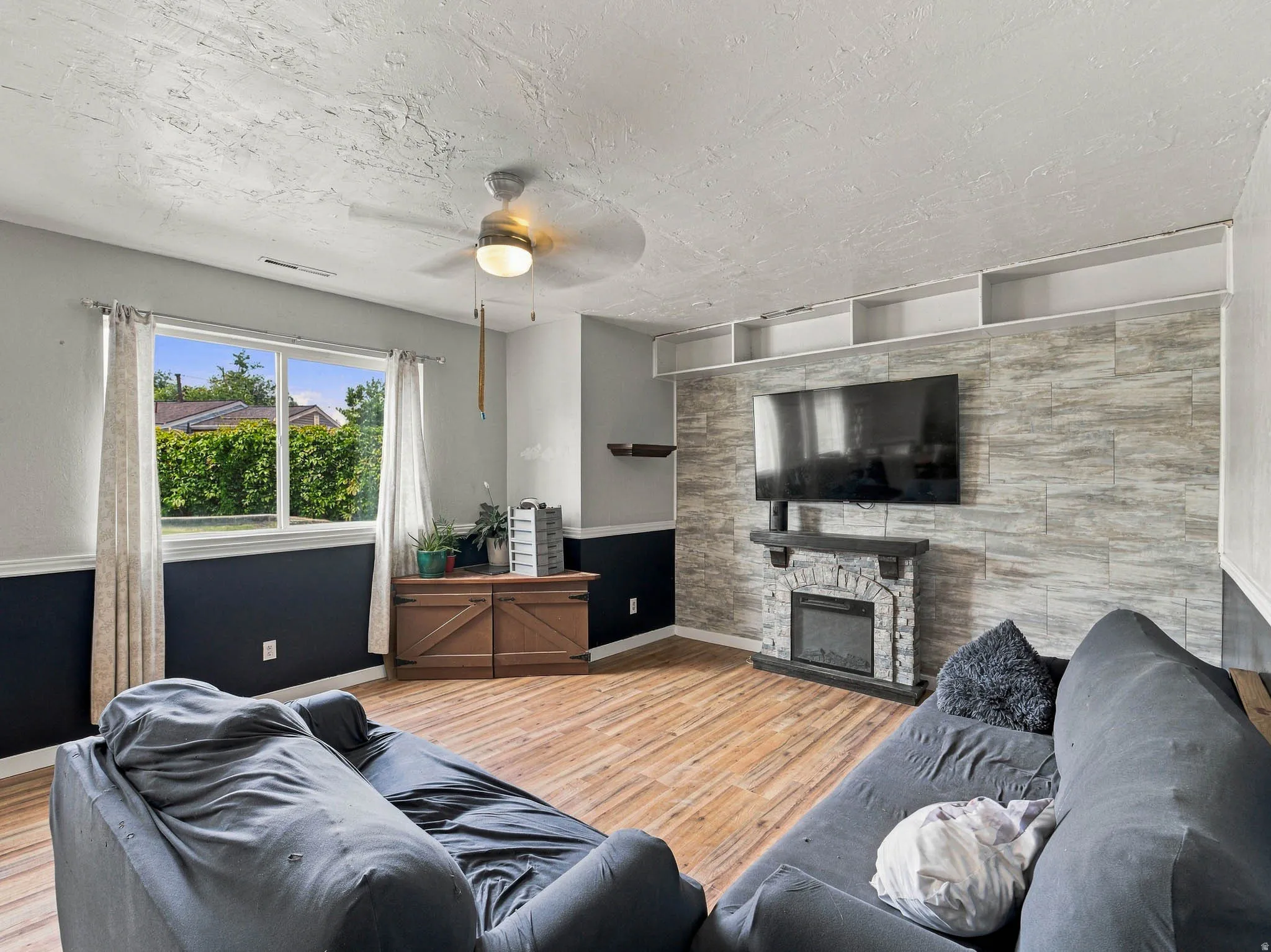 Wood-finish flooring, built-in electric fireplace with stone surround, wall-mounted shelving, ceiling fan, and large window