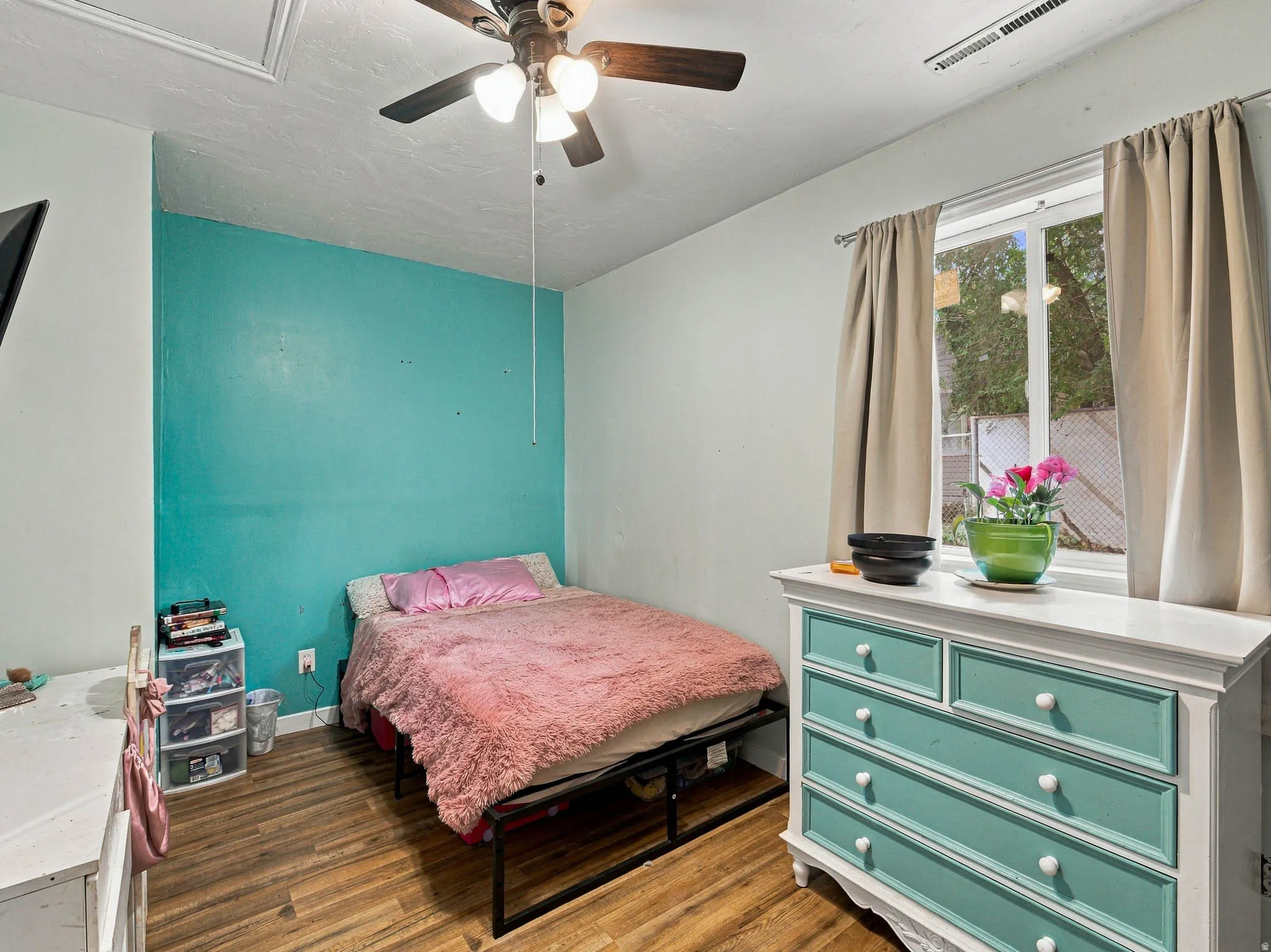 Wood-finish flooring, ceiling fan with integrated lighting, a double-hung window, and an accent wall