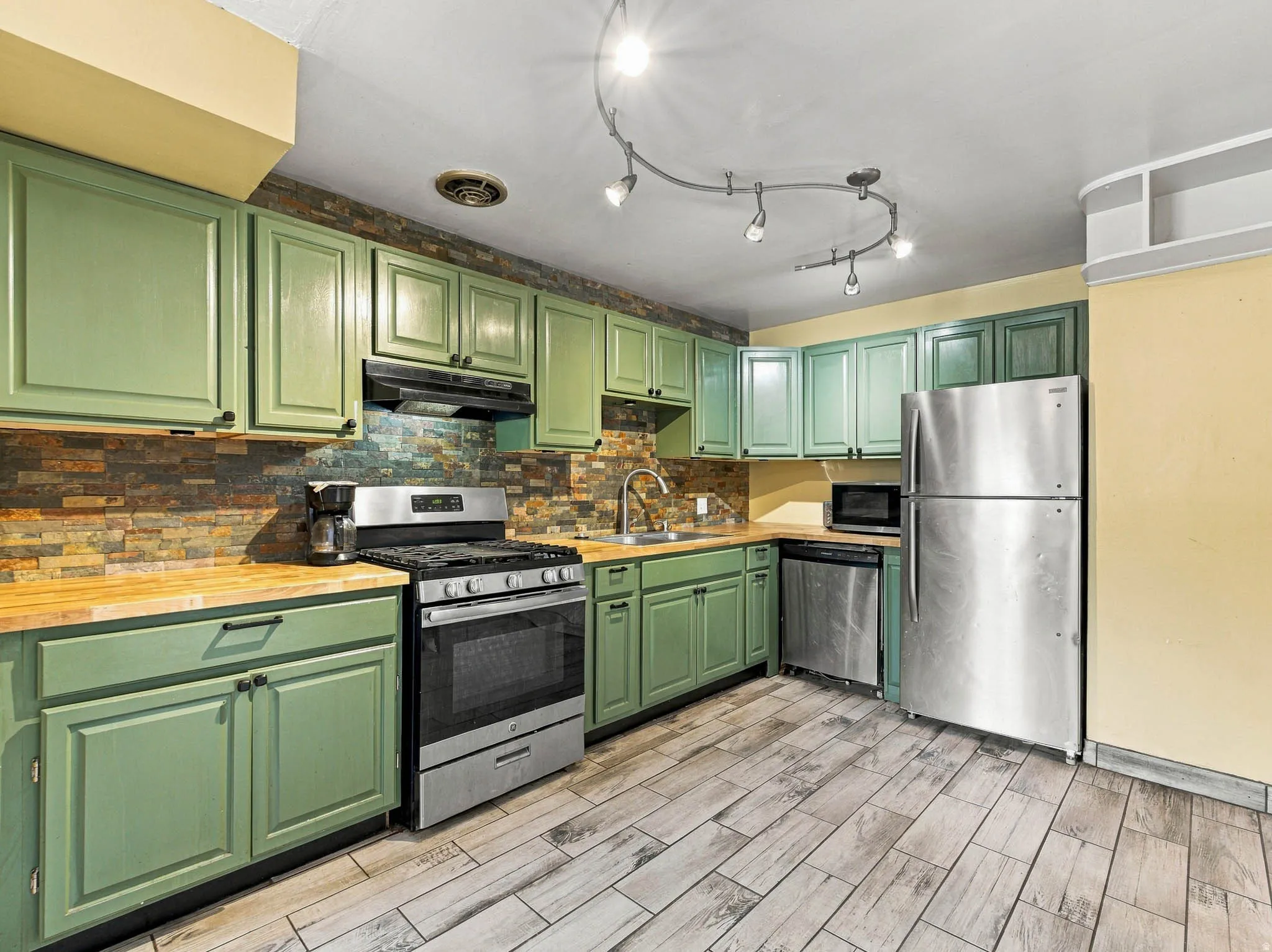Kitchen featuring wood-finish flooring, green cabinetry, stacked stone backsplash, stainless steel appliances, and track lighting