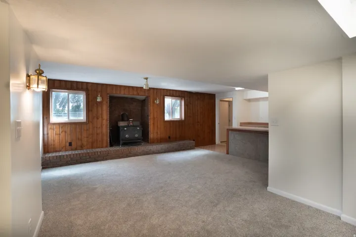 Spacious room featuring wood paneling, a brick-surround fireplace with a stove insert, two windows, and light-toned carpeting