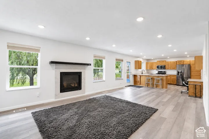 Open concept living area with light wood-finish flooring, a stone-surround fireplace, recessed lighting, and a kitchen featuring wood cabinetry and stainless steel appliances