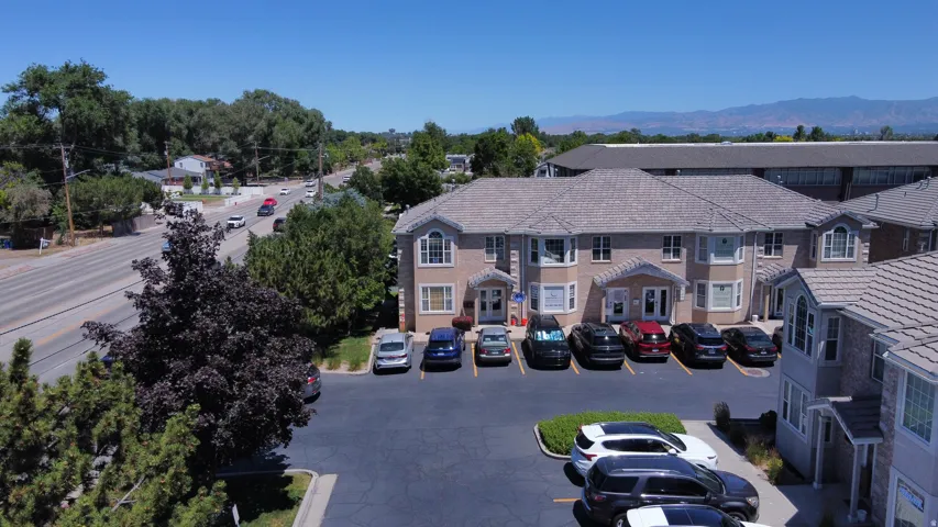 Paved parking lot with marked spaces, bordered by mature landscaping and a two-story building with multiple bay windows and a gray tile roof