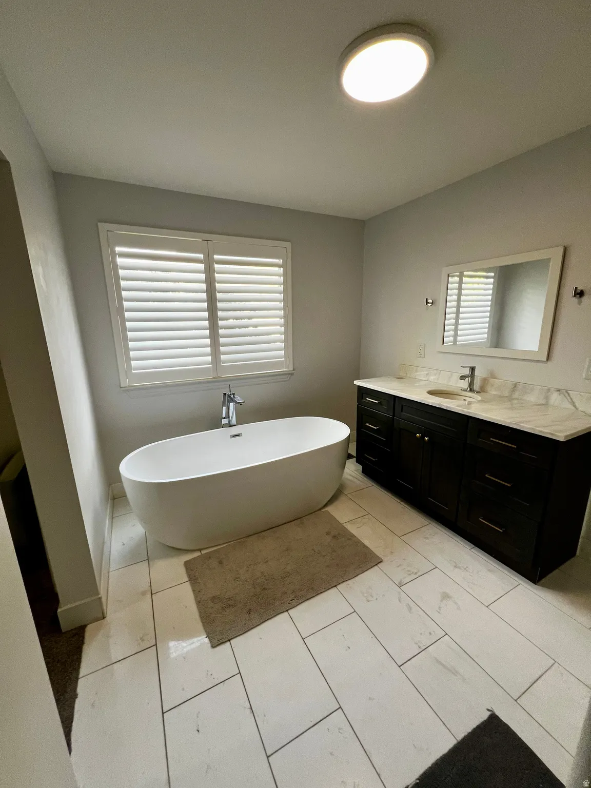 Freestanding soaking tub with floor-mounted faucet, dark wood-finish vanity with countertop, rectangular undermount sink, and tiled flooring