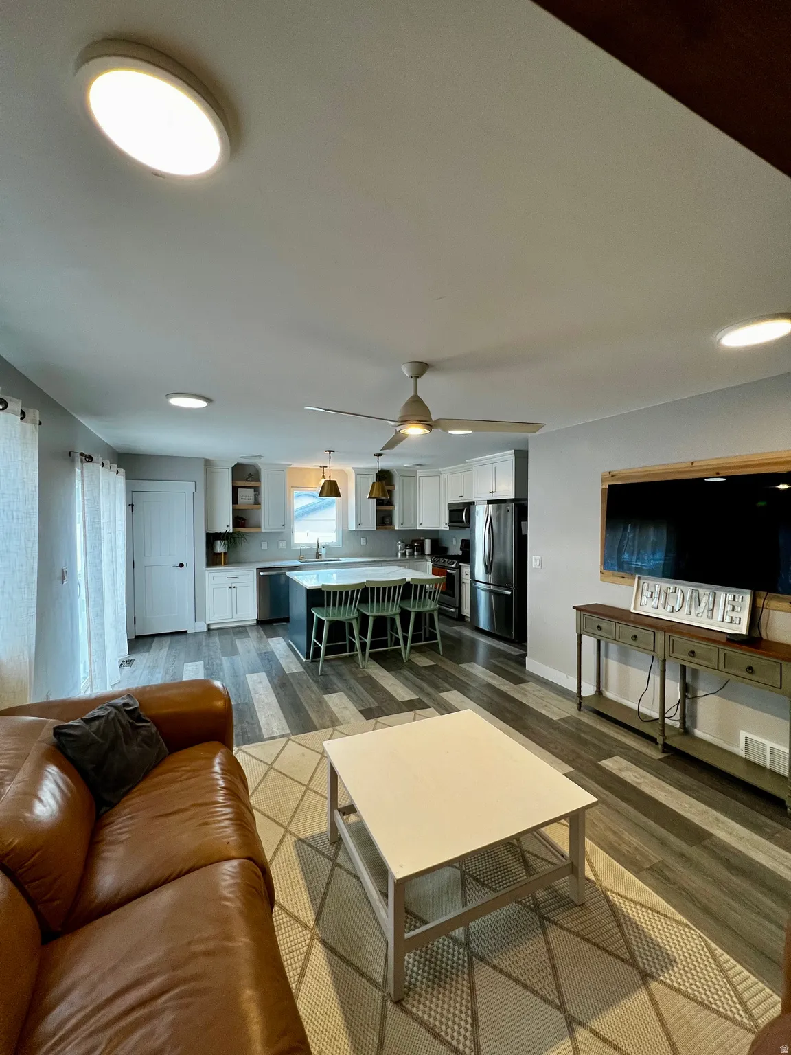 Open-concept living space with wood-finish flooring, white cabinetry, a kitchen island, pendant lighting, and stainless steel appliances